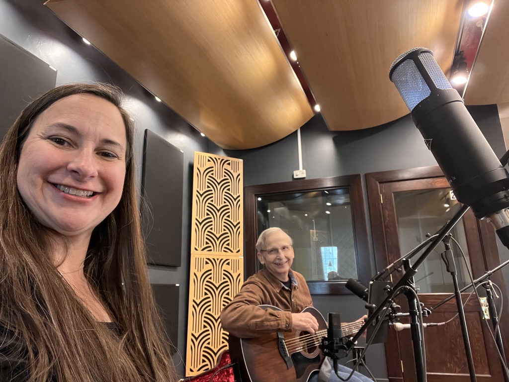 Jess smiling in the studio with Keith on guitar behind her