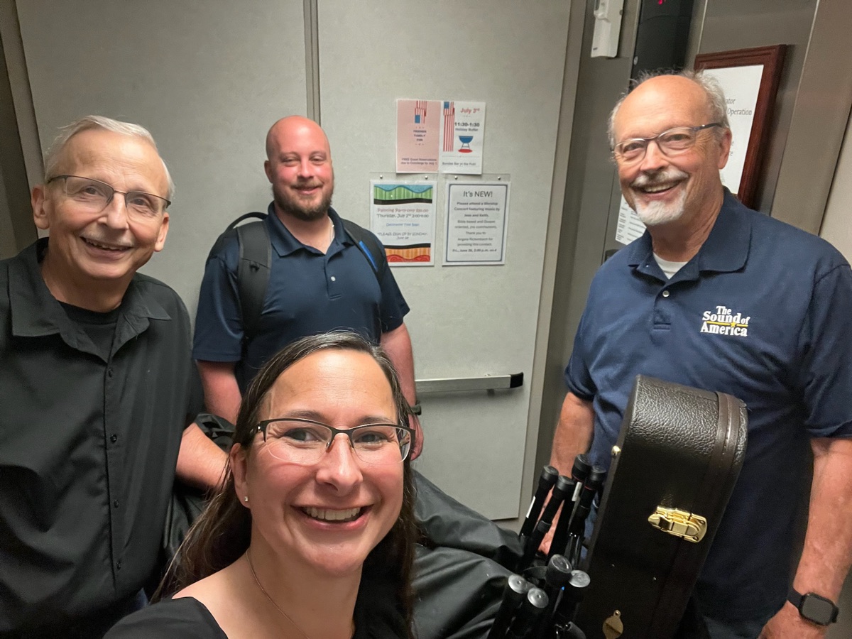 Four team members smiling in an elevator with a guitar case